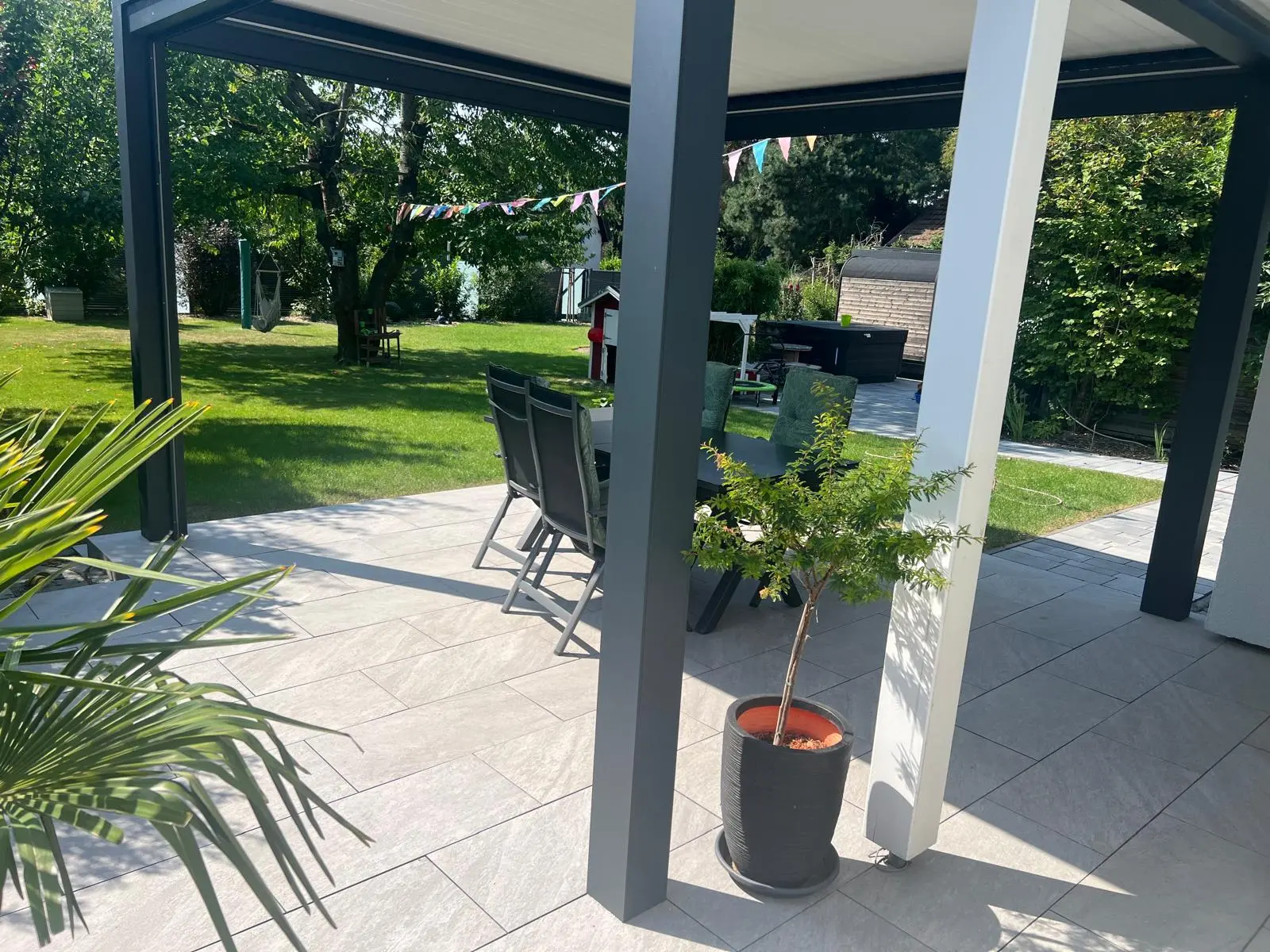 Terrasse mit hellen Platten unter einer dunklen Überdachung, Blick in den Garten.