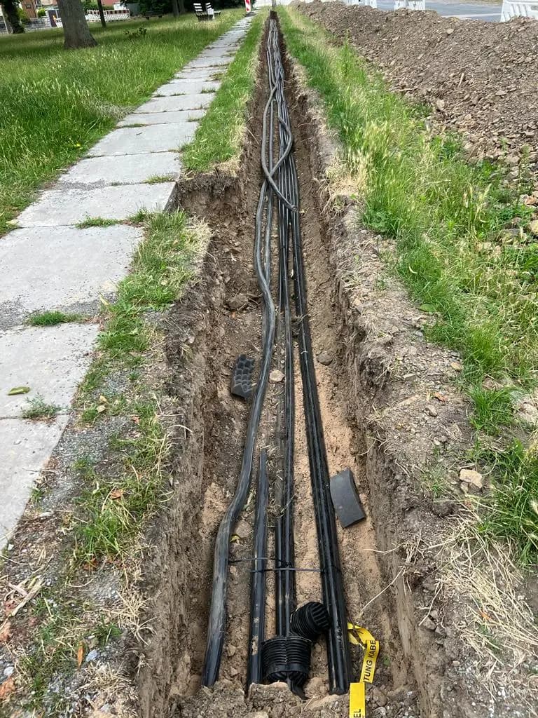 Langer offener Leitungsgraben neben einem Plattenweg, darin mehrere schwarze Schutzrohre.