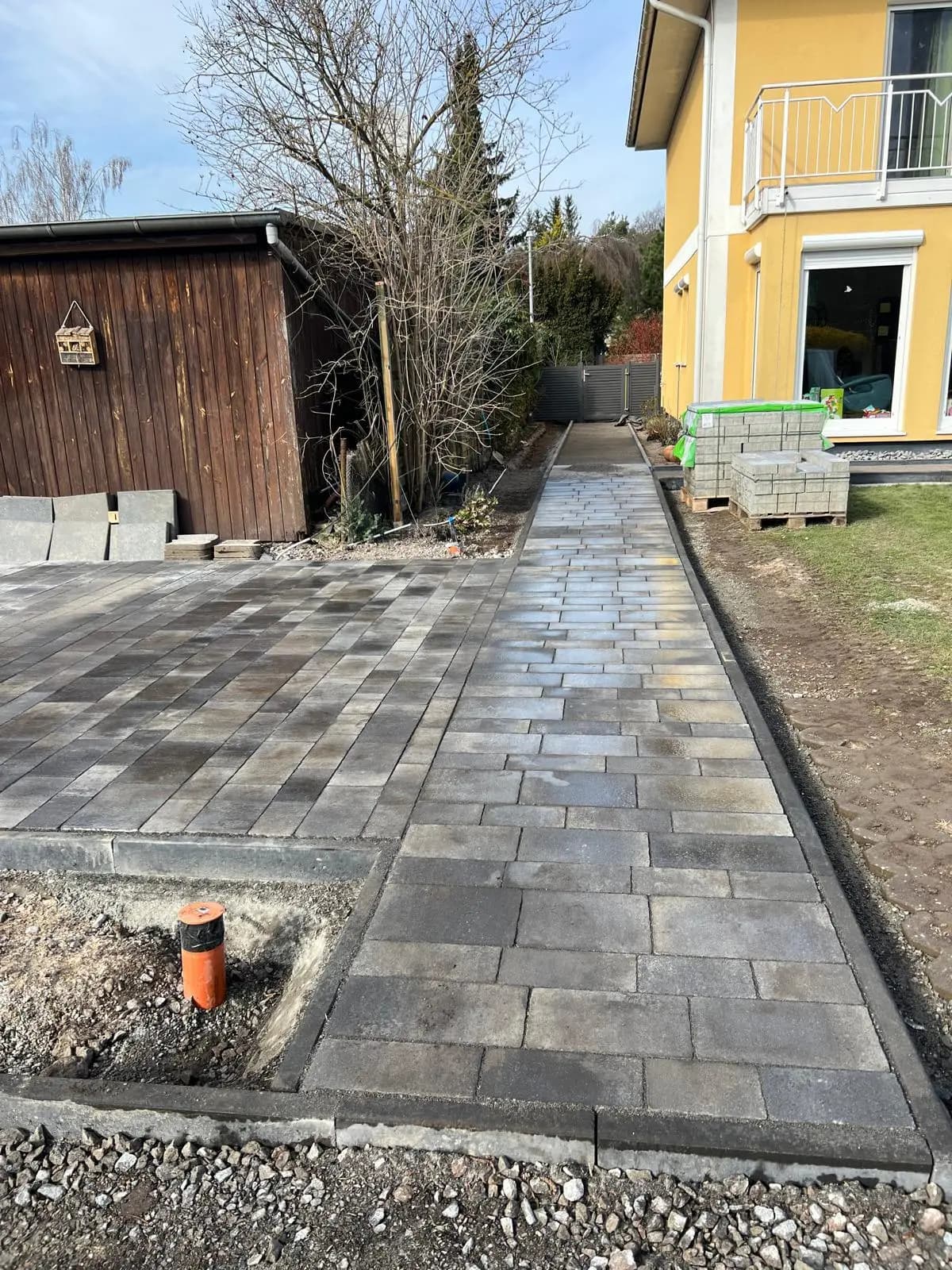 Fertig gepflasterter Weg mit dunkler Einfassung zwischen Holzschuppen und gelbem Haus.